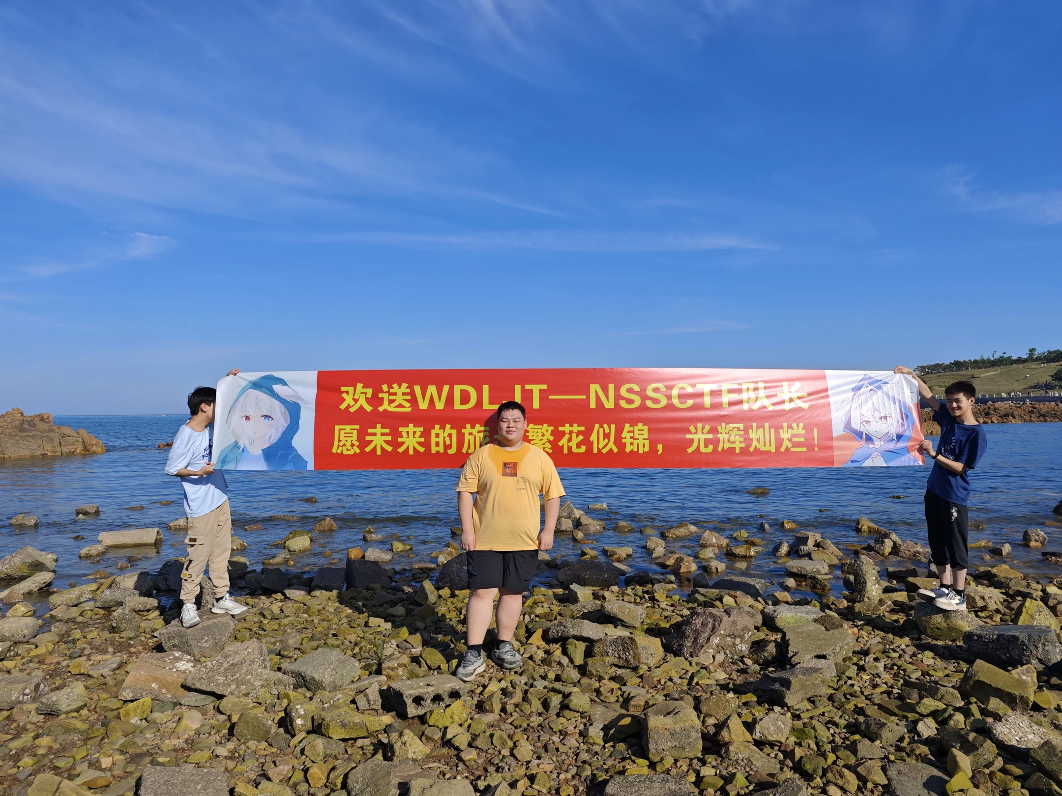 三位队员站在海边岩石上,举着写有欢送和祝福文字的横幅